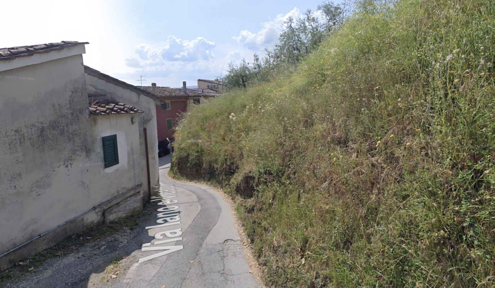 Via Iano e Germinaia: la strada curva stretta tra un edificio e una scarpata alta coperta d'erba, senza muro di sostegno.